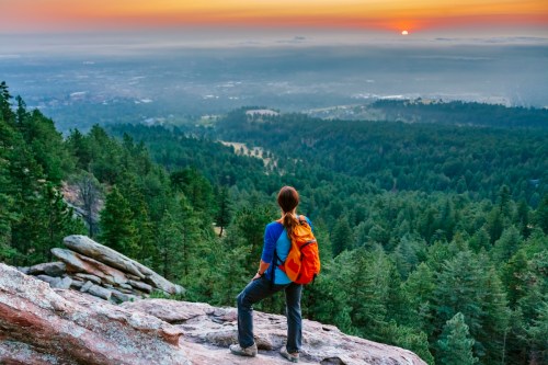 A hiker with a backpack gazes at a sunset over a lush forested valley from a rocky viewpoint.
