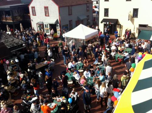 A bustling outdoor market scene with large crowds, tables, and colorful balloons in a festive atmosphere.