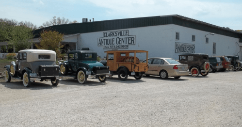 A row of vintage cars parked outside the Clarksville Antique Center, with a large sign on the building in the background.