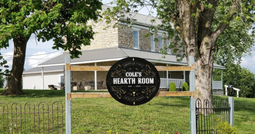 Sign for Cole's Hearth Room in front of a stone building, surrounded by greenery and a well-maintained lawn.