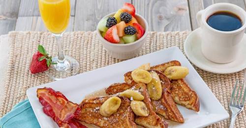 A plate of French toast topped with bananas, served with bacon, fruit salad, a mimosa, and coffee.
