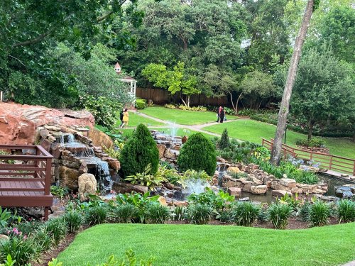 A serene garden with a waterfall, lush greenery, and a wooden bridge, surrounded by trees and colorful flowers.