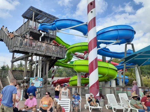 A water park scene featuring colorful water slides, a wooden structure, and visitors relaxing on lounge chairs.