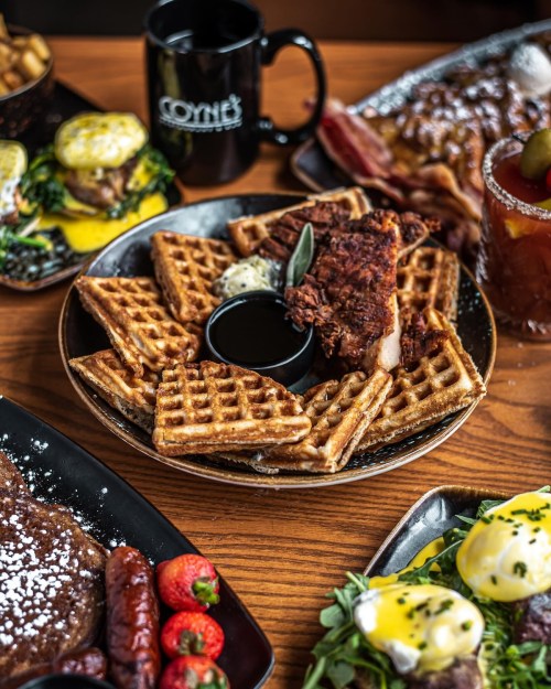 A plate of golden waffles with fried chicken, served with a small cup of syrup, alongside a coffee mug and other dishes.
