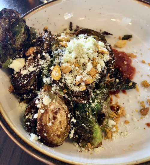A bowl of roasted Brussels sprouts topped with crumbled cheese, crushed nuts, and a drizzle of sauce.