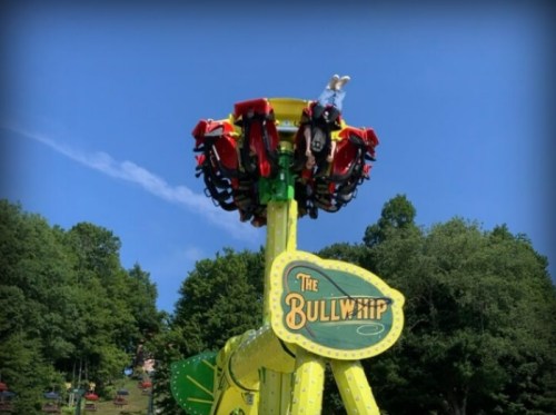 A colorful amusement park ride called "The Bullwhip" with riders suspended high against a blue sky and green trees.