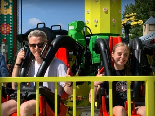 A man and a girl give thumbs up while seated on a colorful amusement park ride, ready for an exciting experience.