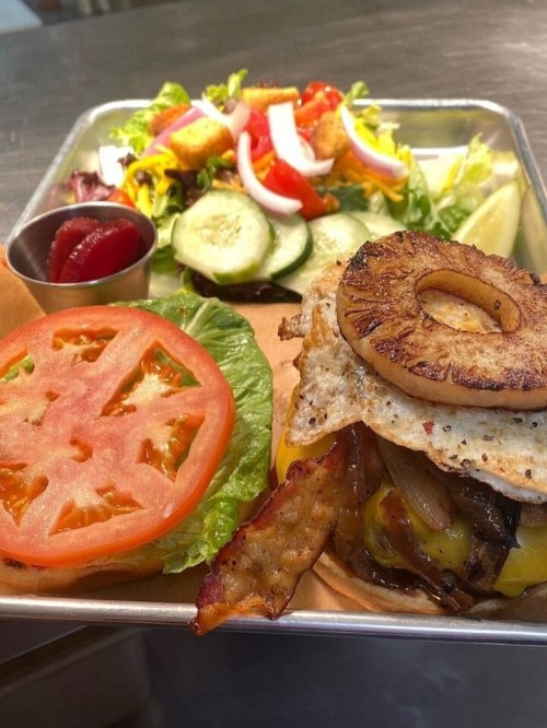 A delicious burger with bacon, cheese, and a fried egg, served with a fresh salad and a side of pickles.