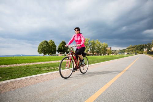 A woman in a pink jacket rides a bicycle along a path with green grass and trees under a cloudy sky.