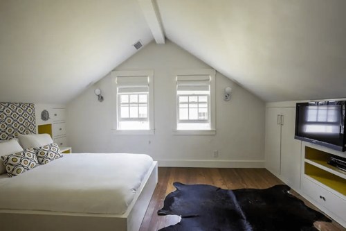 Bright attic bedroom with a bed, patterned headboard, two windows, and a TV on a white cabinet. Wood flooring and a rug.