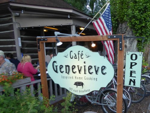 Sign for Café Genevieve with "Open" banner, surrounded by greenery and bicycles, in a rustic setting. American flag visible.