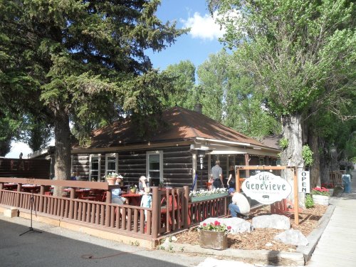 A rustic log cabin café with outdoor seating, surrounded by trees and flowers, featuring a sign that says "Café Geneviève."
