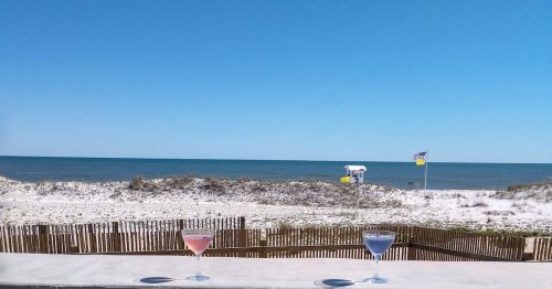Two colorful cocktails sit on a table overlooking a sandy beach and calm ocean under a clear blue sky.