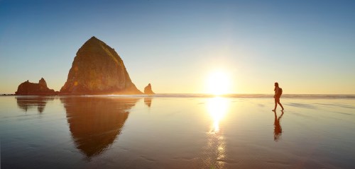 A person walks along a reflective beach at sunset, with a large rock formation silhouetted against the colorful sky.