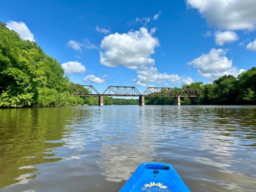 A view from a kayak on a calm river, featuring a bridge and lush green trees under a blue sky with fluffy clouds.