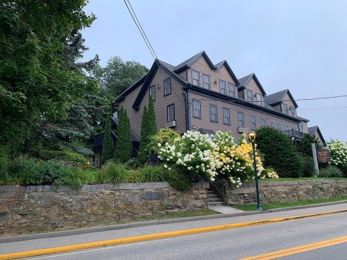 A large, dark wooden building surrounded by lush greenery and blooming white flowers, set along a quiet street.