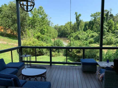 A serene view from a porch overlooking a lush green landscape and a calm river. Comfortable seating is visible.