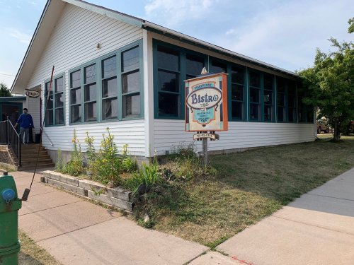 A white building with green windows and a sign reading "Bistro" in front, surrounded by grass and plants.