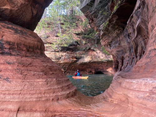 A person kayaking through a narrow rock formation with vibrant red and green hues in a serene waterway.