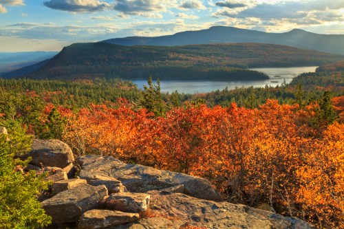 A scenic view of a valley with vibrant autumn foliage, a lake, and mountains under a cloudy sky.