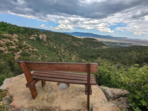 A wooden bench overlooks a lush green valley and distant mountains under a cloudy sky.