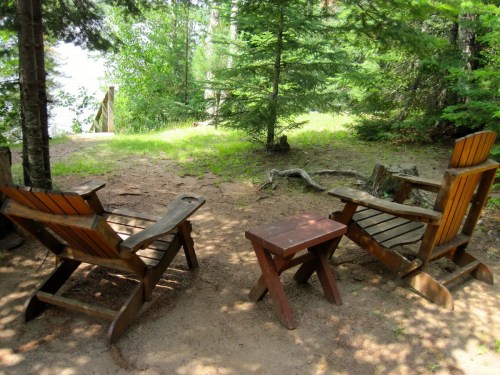 Two wooden chairs and a small table sit on a sandy patch surrounded by trees, overlooking a serene lake.