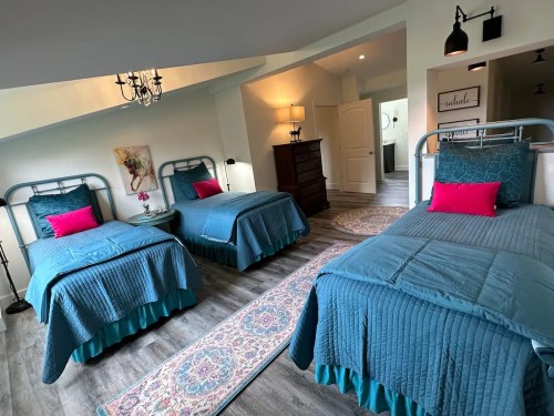 A cozy bedroom with three twin beds, teal bedding, and a stylish rug, featuring soft lighting and a warm atmosphere.