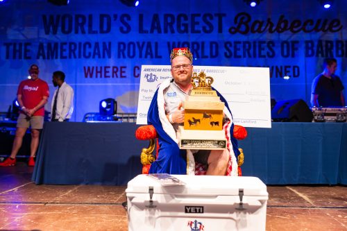 A man in a crown and robe sits on a throne, holding a trophy and a large check at a barbecue competition event.