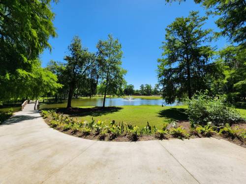 A serene park scene featuring a pond, fountain, and lush greenery under a clear blue sky.