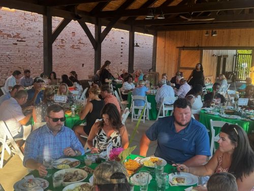 A lively gathering of people dining at tables in a rustic venue, enjoying food and conversation.