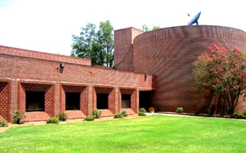 A brick building with a cylindrical structure, surrounded by green grass and trees under a clear blue sky.