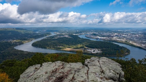 A panoramic view of a winding river surrounded by lush greenery and distant hills under a partly cloudy sky.