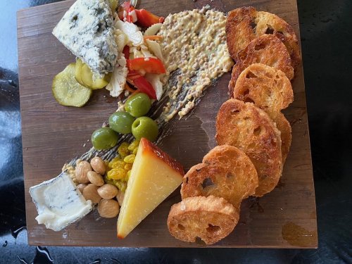 A wooden platter featuring assorted cheeses, olives, pickles, nuts, and crispy bread slices.