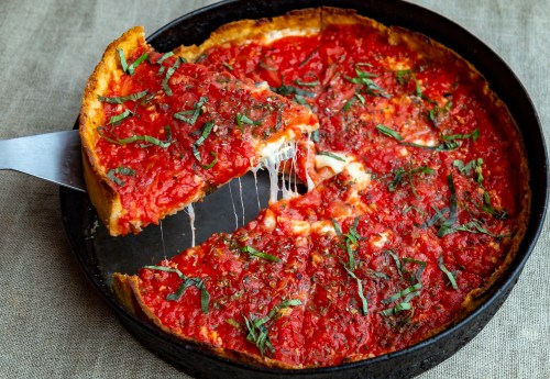 A deep-dish pizza with a thick crust, topped with tomato sauce, cheese, and fresh basil, with a slice being lifted.