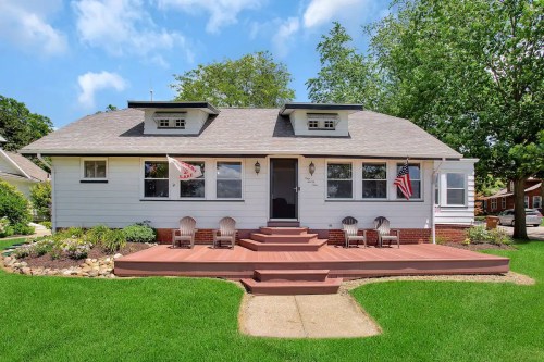 A charming white house with a front porch, surrounded by green grass and trees, featuring American flags and seating.