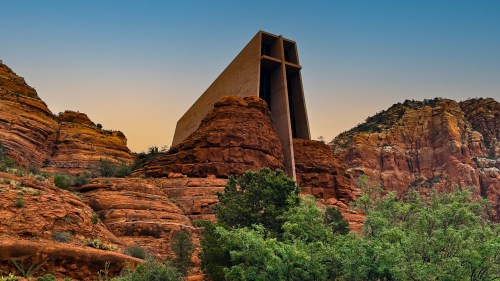 A modern building with a cross design, set against red rock formations and greenery under a clear sky.