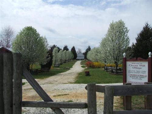 A scenic pathway lined with flowering trees leads to Clover Hill Village, with a welcoming sign at the entrance.