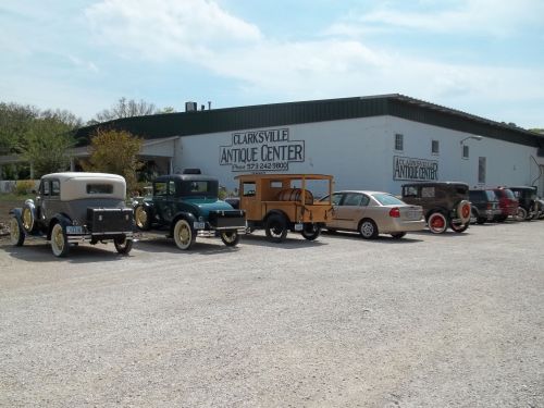 A row of vintage cars parked outside the Clarksville Antique Center, with a large building in the background.