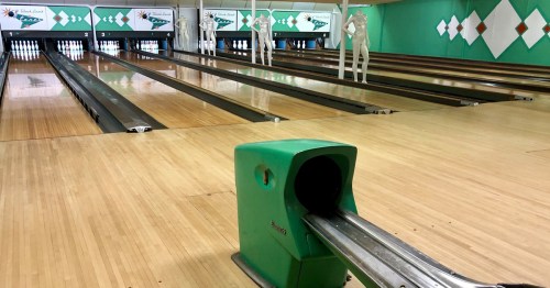 A bowling alley with green ball return, wooden lanes, and white bowling pins set up at the end.