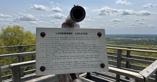 A viewpoint with a landmark locator sign and a telescope overlooking a scenic landscape under a cloudy sky.