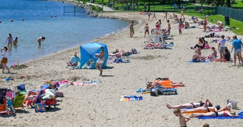 A busy beach scene with people sunbathing, swimming, and relaxing on the sand near the water.