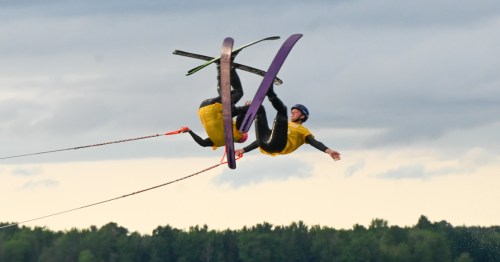 A water skier performs a mid-air trick, soaring above the water with skis and a tow rope against a cloudy sky.