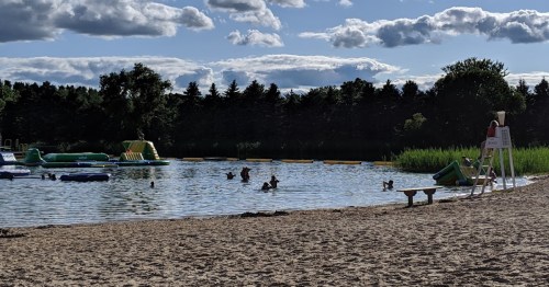 A sandy beach with people swimming in a lake, inflatable toys, and a lifeguard on duty under a partly cloudy sky.