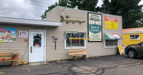 Exterior of Scottie's Bait & Tackle shop, featuring a large fish sign, open banner, and colorful advertisements.