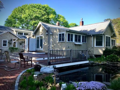 A charming cottage with a wooden deck, surrounded by greenery and a small pond in the foreground.