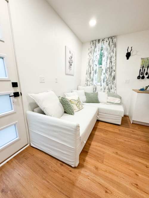 Cozy living space with a white sofa, green accents, and a bright window, featuring light wood flooring and decorative curtains.
