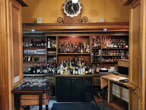 A wooden bar with shelves filled with various bottles, glasses, and a clock on the wall.