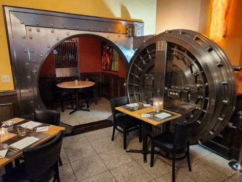A restaurant interior featuring a large, metallic vault door and tables set for dining.