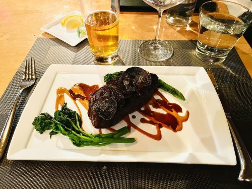 A plated steak with sauce, asparagus, and broccolini, accompanied by a beer and glasses of water on a table.
