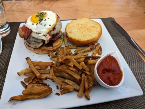 A burger with a fried egg, bacon, and cheese on a plate, accompanied by fries and a small bowl of ketchup.